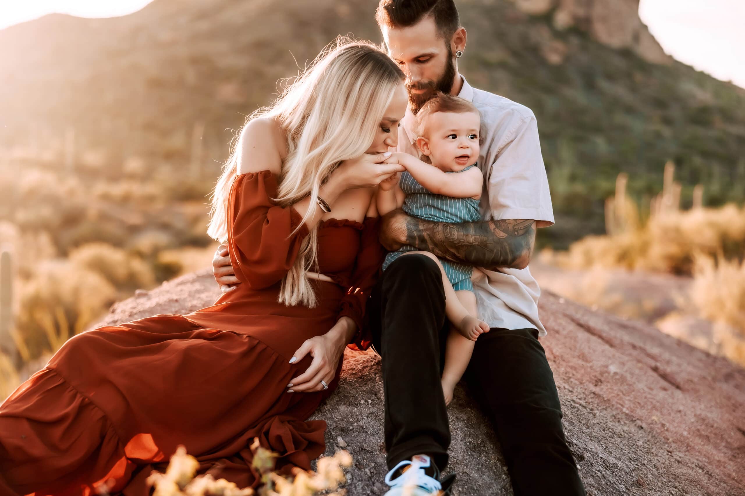 Mom kisses baby boy's hand as he sits in his dad's lap for their family session.